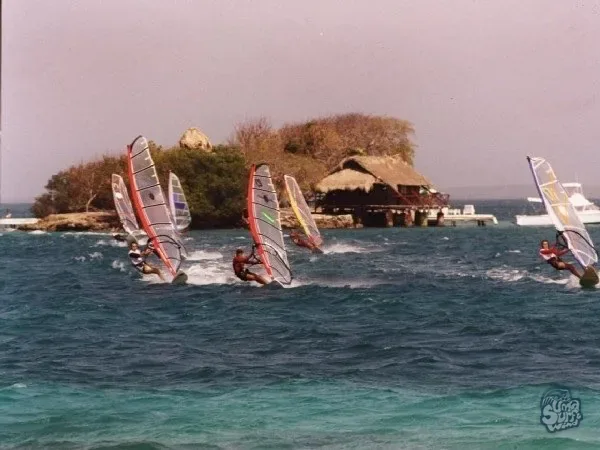 Flota de windsurfistas navegando junto a un islote con casa de palma