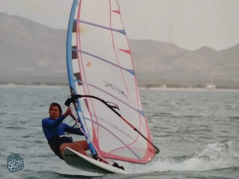 Windsurfista planeando con vela rosa y azul frente a la costa